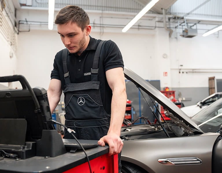 Kunzmann Werkstatt-Monteur an seinem Laptop vor einem Mercedes-Benz in der Werkstatt Kunzmann Werkstatt-Monteur an seinem Laptop vor einem Mercedes-Benz in der Werkstatt